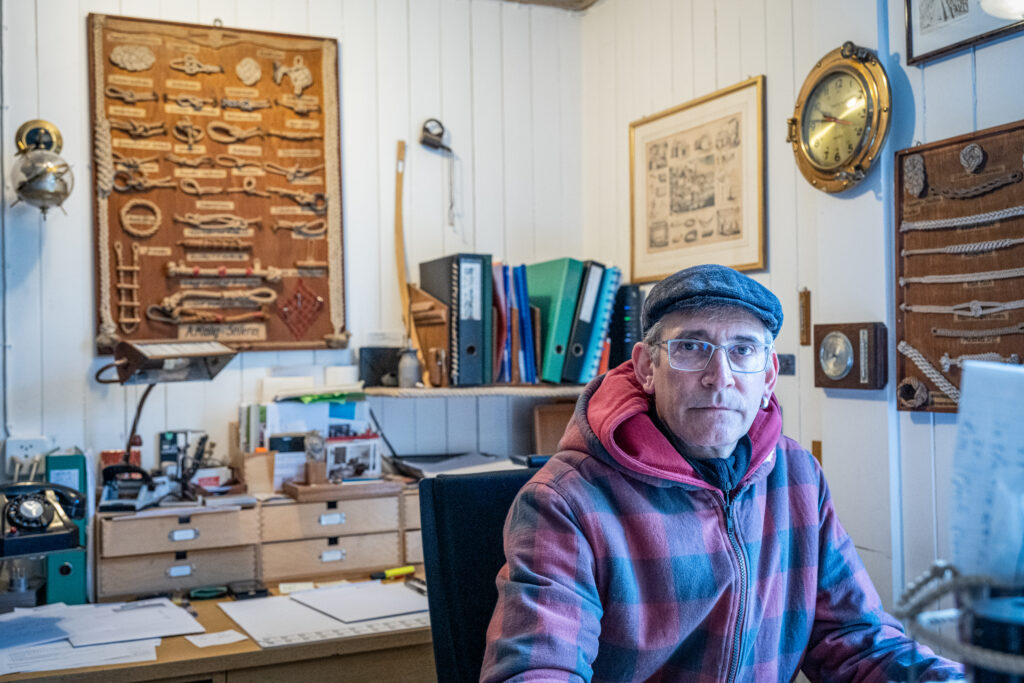 Inhaber der Seilerei Kislig, Martin Benz im B&uuml;ro. Im B&uuml;ro h&auml;ngen neben einer alten Seemannsuhr, verschieden Schautafeln auf denen Seile fixiert sind, die verschiedene Knoten und Flechtarten von Seilen zeigen.